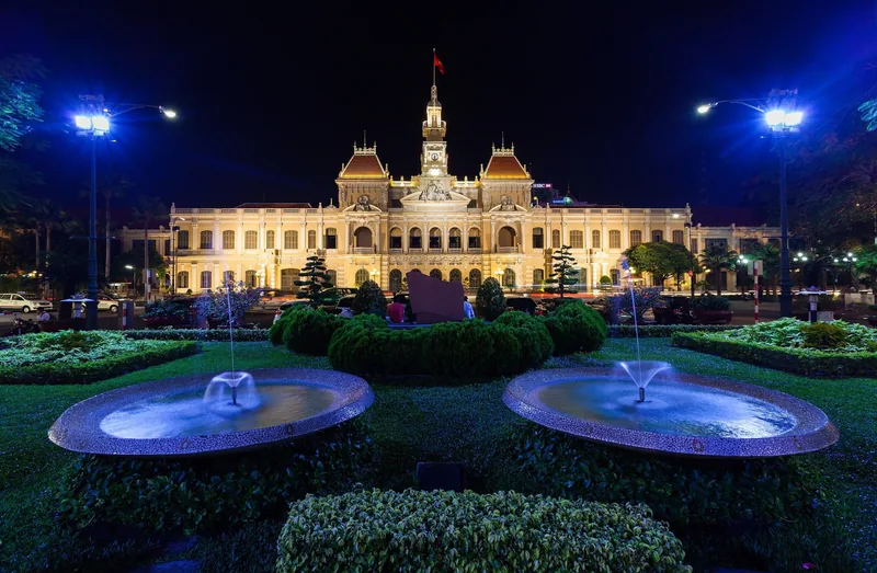 Tòa nhà Ủy ban Nhân dân Thành phố Hồ Chí Minh rực sáng về đêm phía sau hai vòi phun nước và khu vườn cắt tỉa gọn gàng.